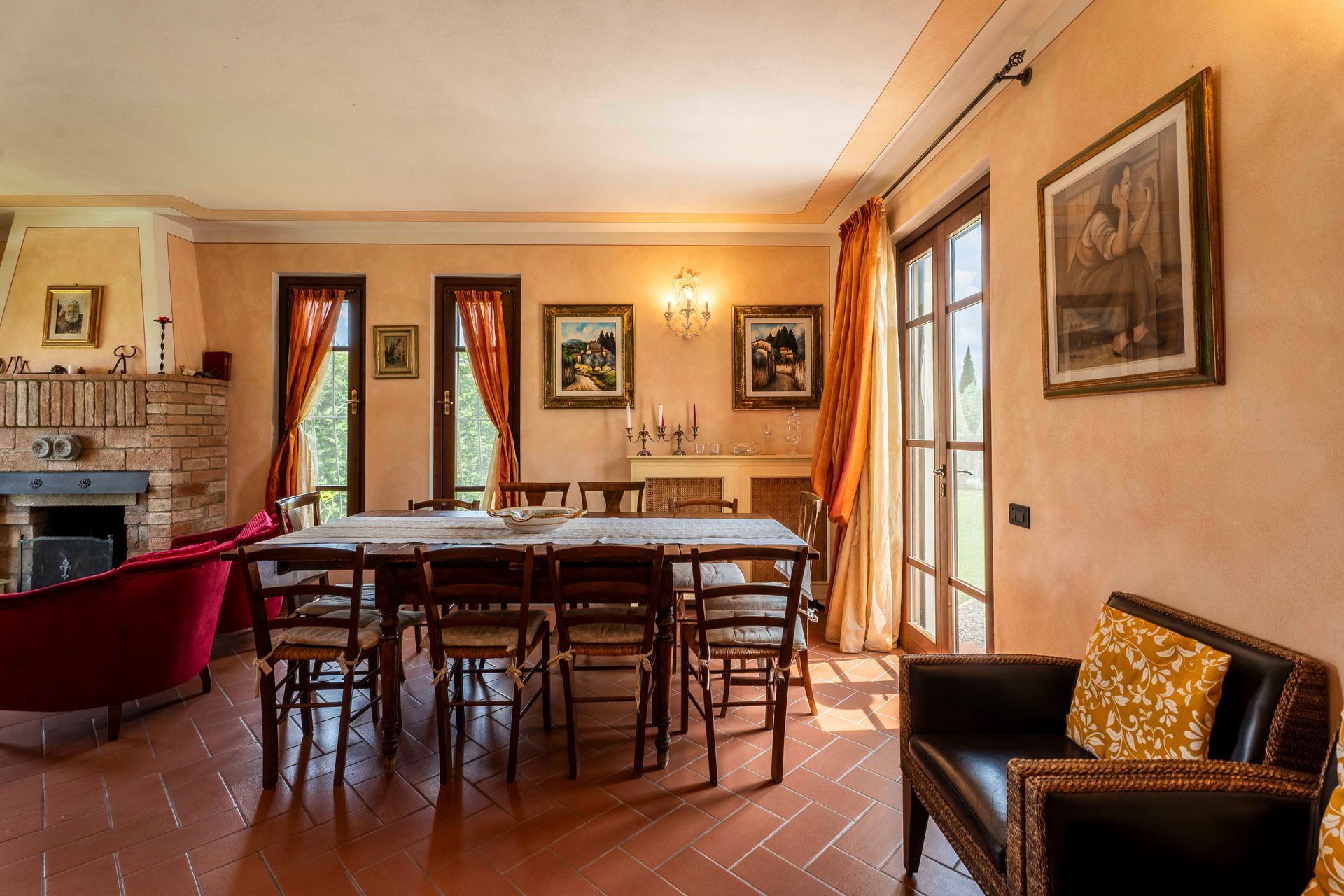 Accogliente sala da pranzo con lungo tavolo, sedie in legno, pavimento in cotto e quadri incorniciati vicino a finestre illuminate dal sole.