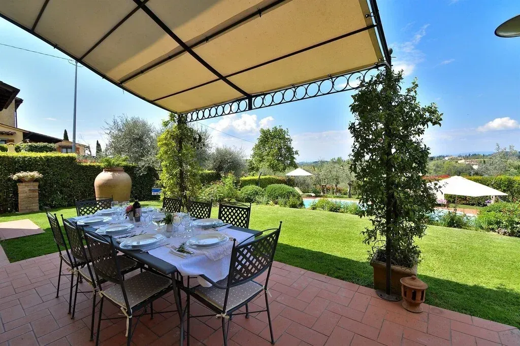 Zona pranzo all'aperto sotto una tenda da sole beige con vista su un giardino verdeggiante e sugli alberi in una giornata di sole.