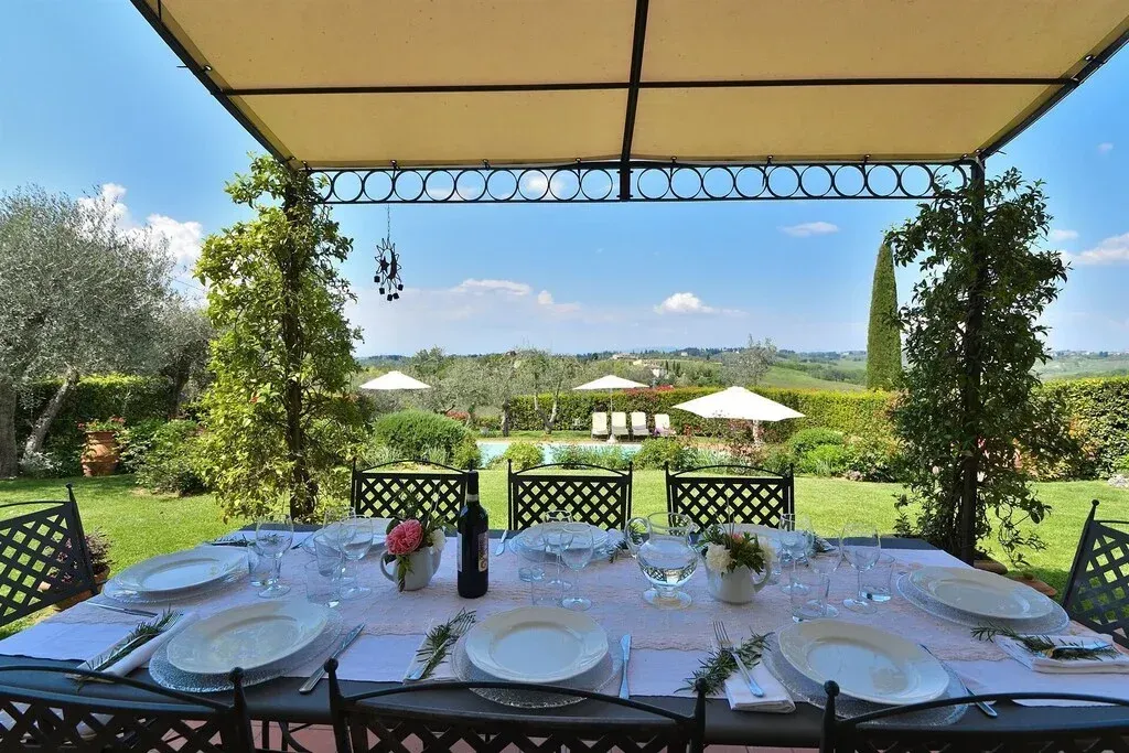 Tavolo da pranzo all'aperto sotto un gazebo con vista su un giardino soleggiato e sulle colline in lontananza.