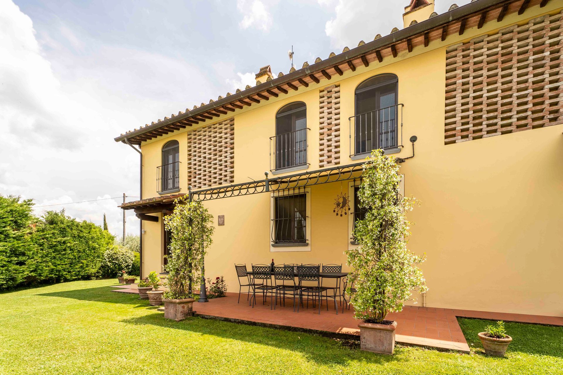 Casa gialla con finestre con persiane, patio ricoperto di viti e prato verde sotto un cielo luminoso