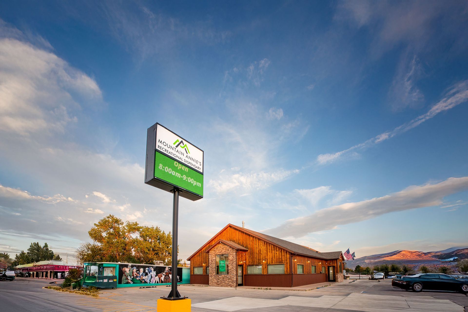 A brown wooden building with a green-and-white sign stands under a cloudy sky near mountains at sunset.