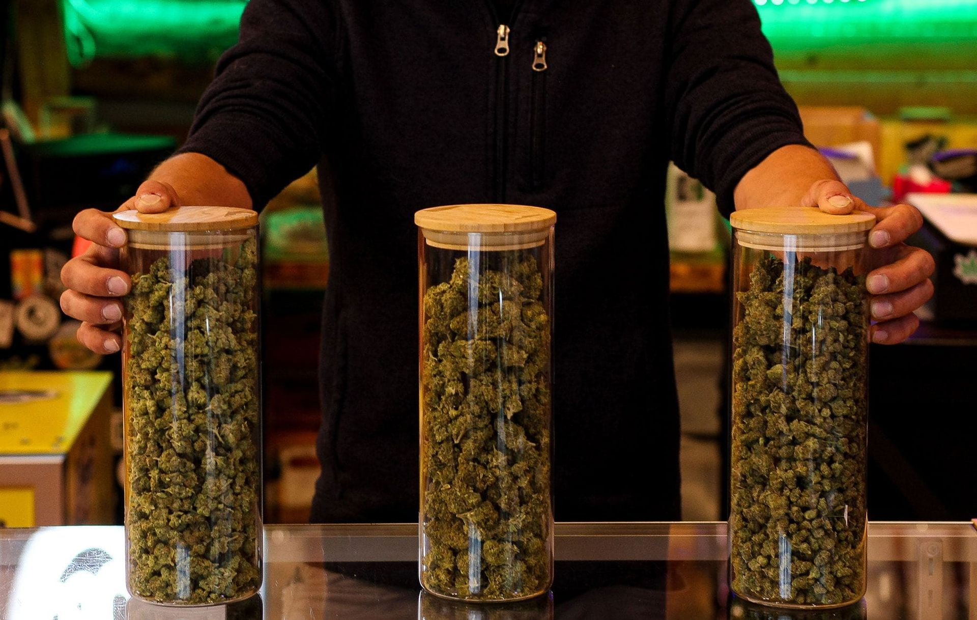A person holds three tall glass jars filled with cannabis flower buds behind a shop counter.