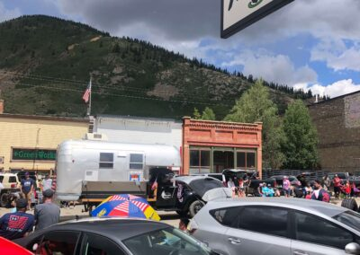 A small vintage silver camper parked on a city street in front of a mountain under a cloudy blue sky.
