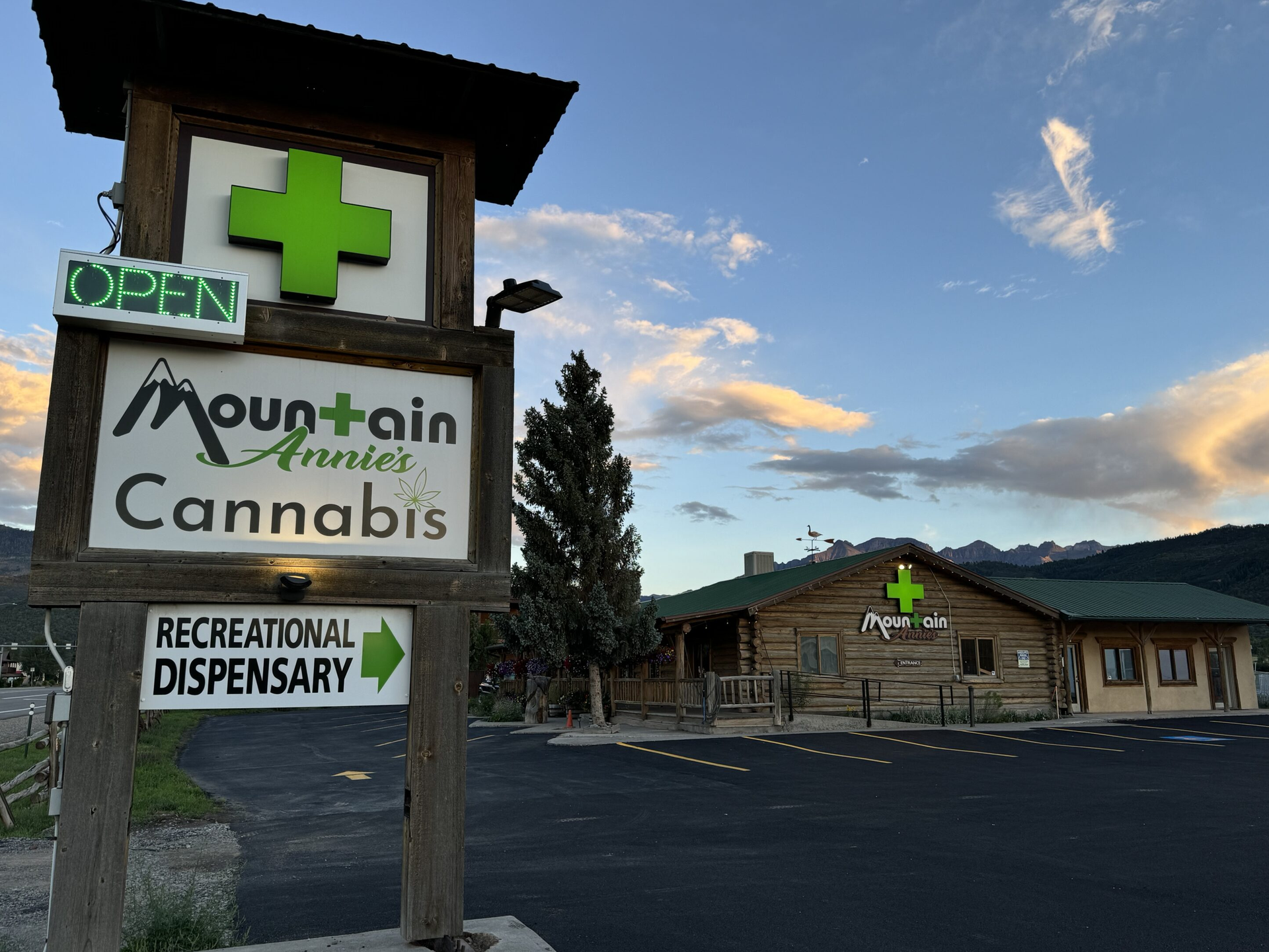 A wooden sign for Mountain Annie’s Cannabis featuring a green cross, standing before a log cabin building at sunset.
