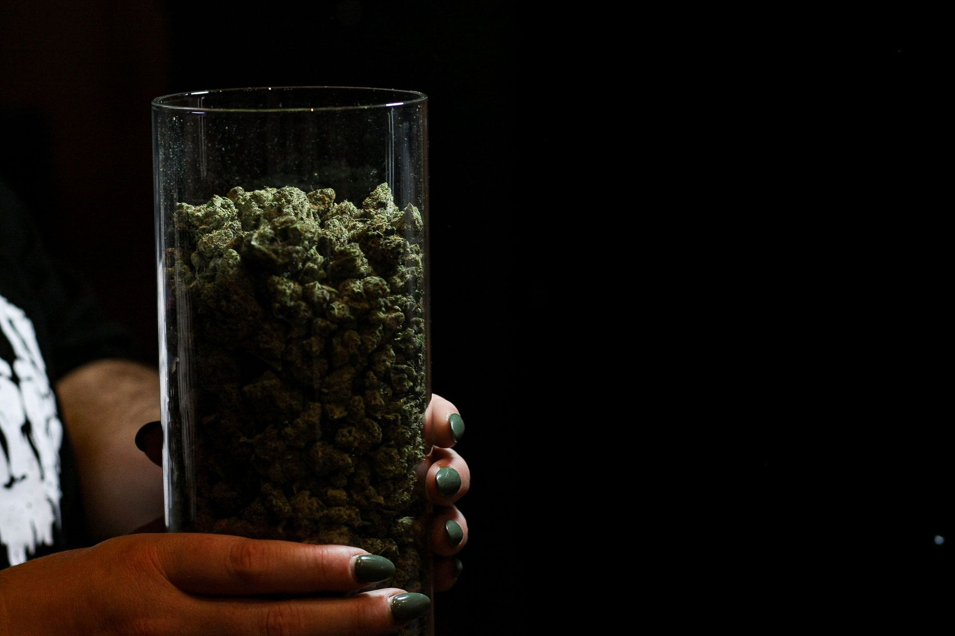 A person holds a tall glass cylinder filled with small, dried green cannabis flower buds against a dark background.