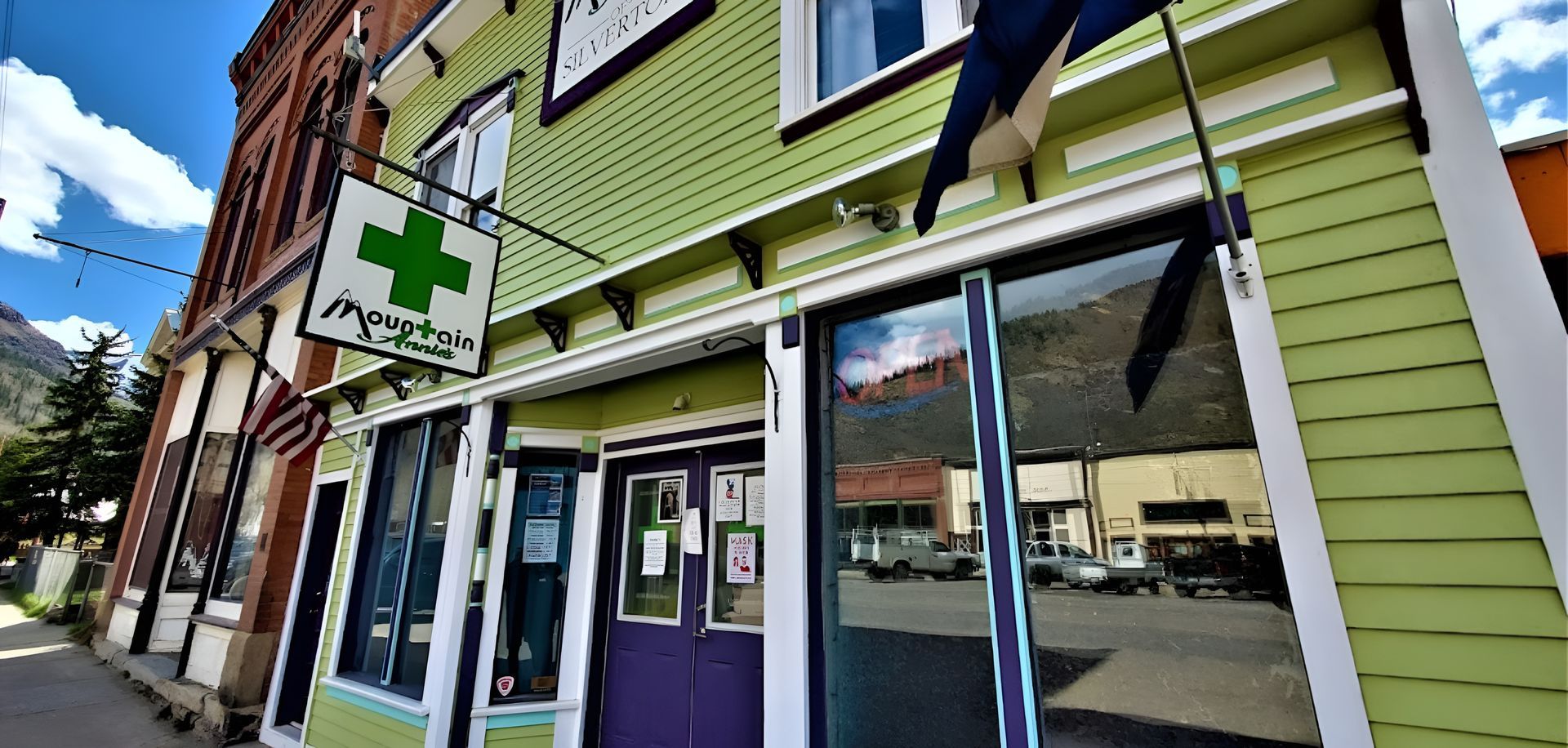 A green, historic storefront in a mountain town, featuring a hanging sign with a green cross and pharmacy branding.