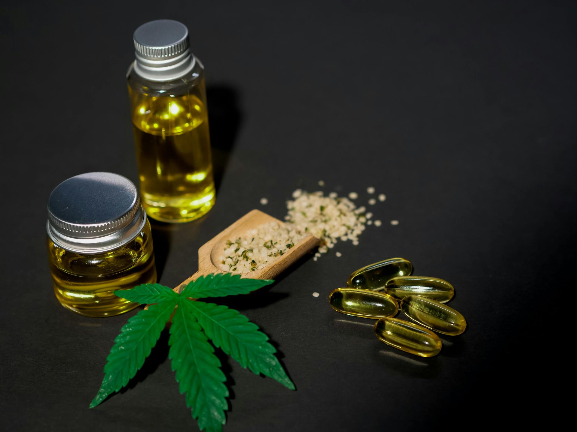 Cannabis oil in bottles, a wooden scoop of hemp seeds, capsules, and a green cannabis leaf on a black background.