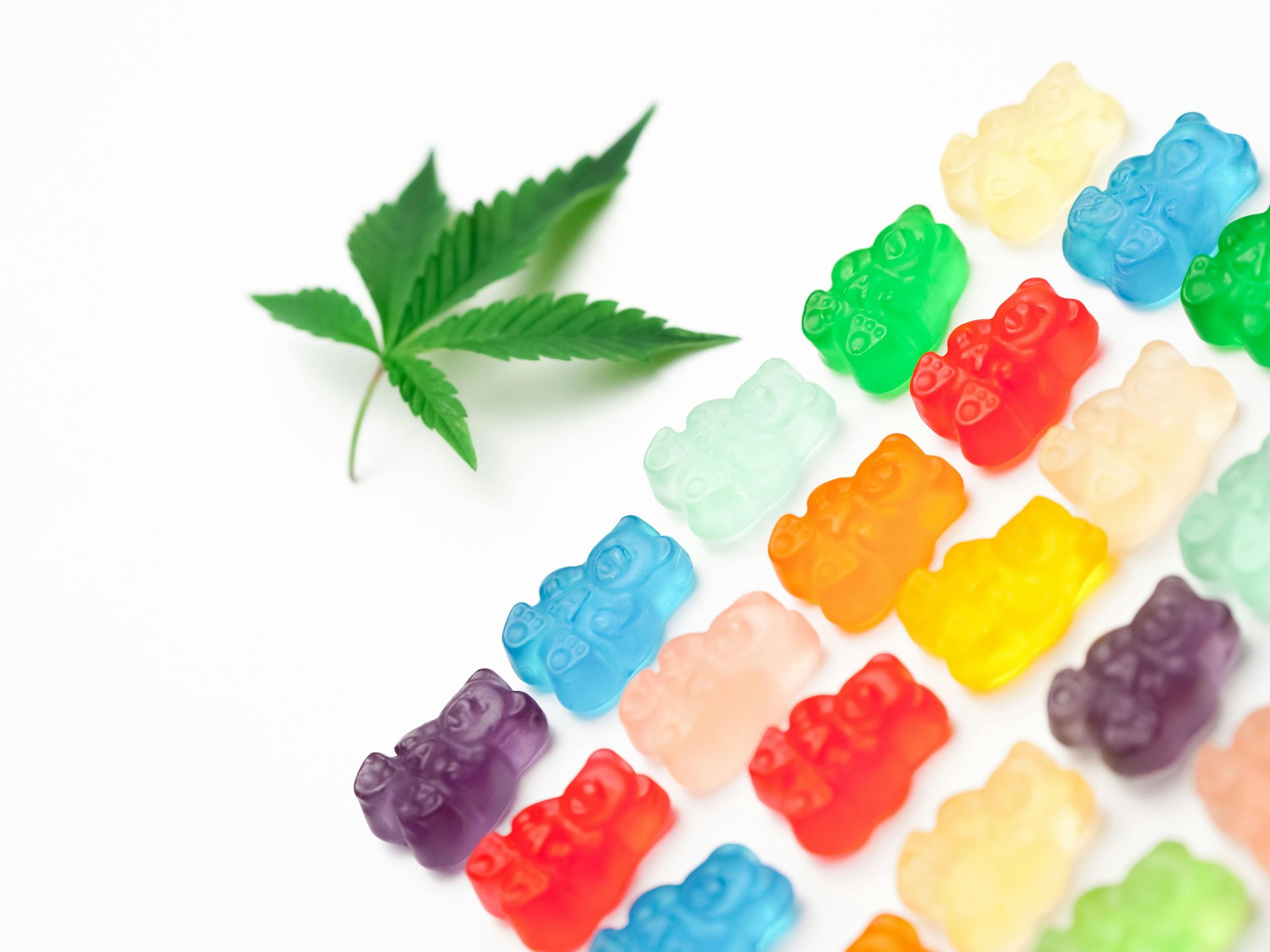 A cannabis leaf positioned next to a diagonal arrangement of multicolored gummy bears on a white background.