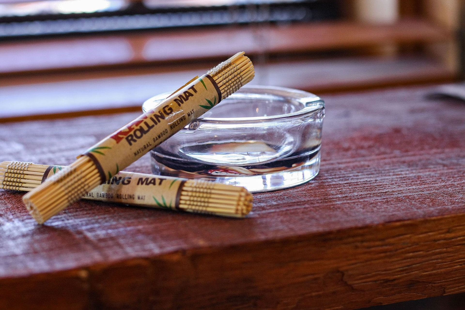 Two bamboo rolling mats resting on a dark wood surface next to a glass ashtray.