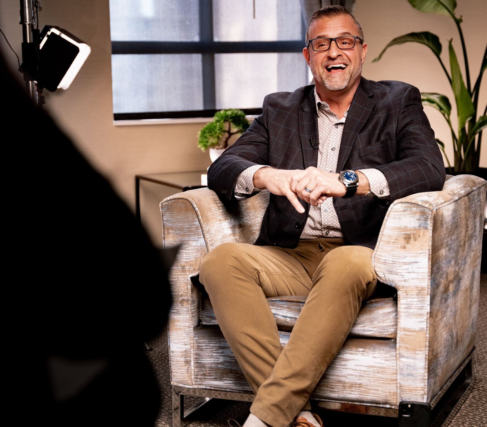 A man in a suit and glasses is sitting in a chair and smiling.