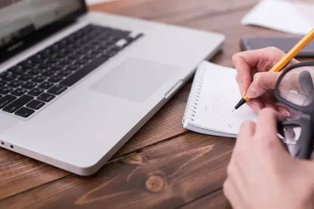 A person is writing in a notebook in front of a laptop computer.