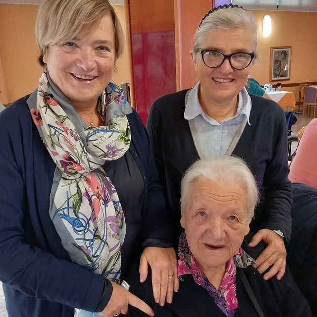 Tre persone sorridenti in posa al chiuso, due delle quali in piedi dietro a un’anziana seduta.