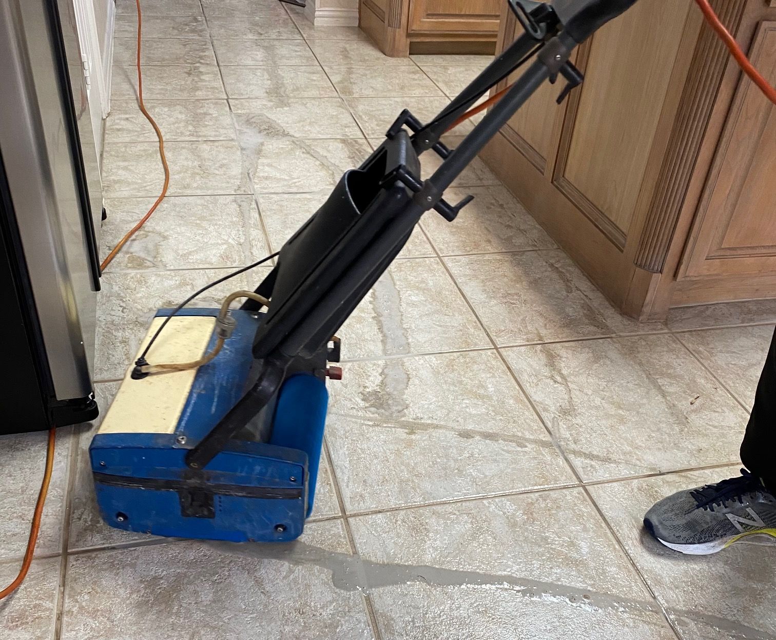 A person using a blue and beige floor cleaning machine on tile floor in a kitchen. A person using a blue and beige floor cleaning machine on tile floor in a kitchen.