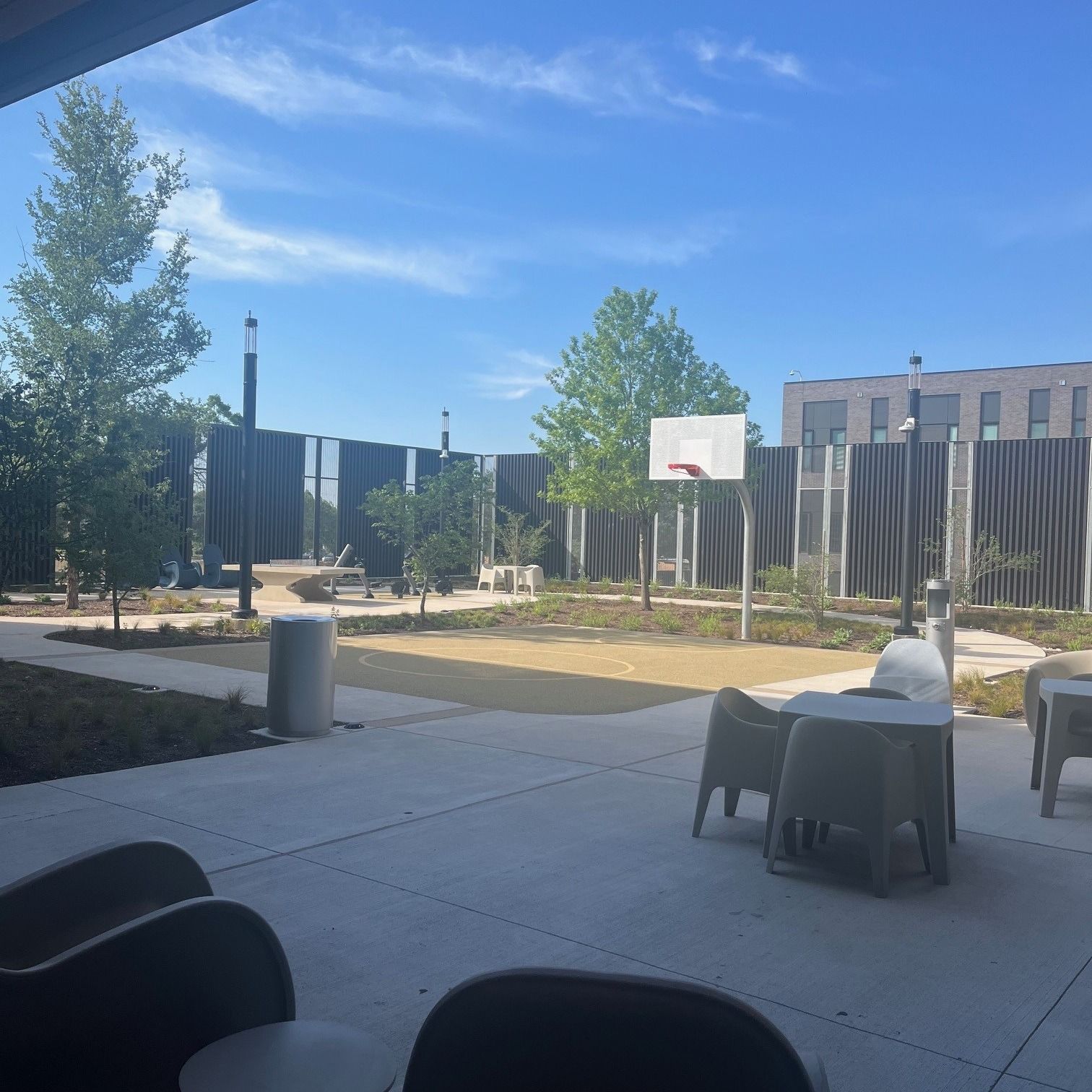 Outdoor patio with tables, chairs, trees, and a basketball hoop beside a modern building under a blue sky