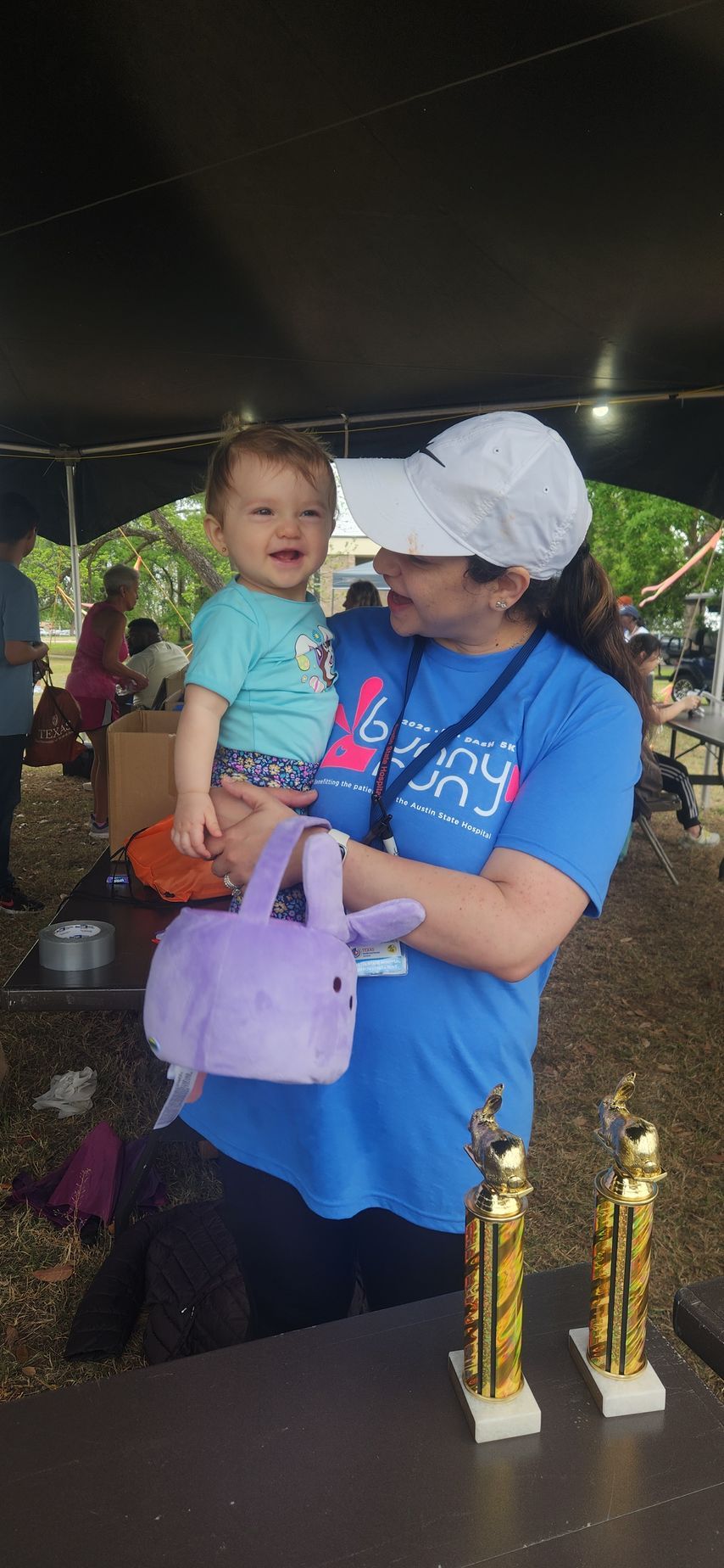 Friends of ASH volunteer holding a child at an outdoor Austin community event with award trophies on display
