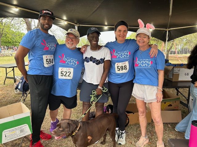 Five runners in blue shirts pose with a brown dog under a canopy at a race event.