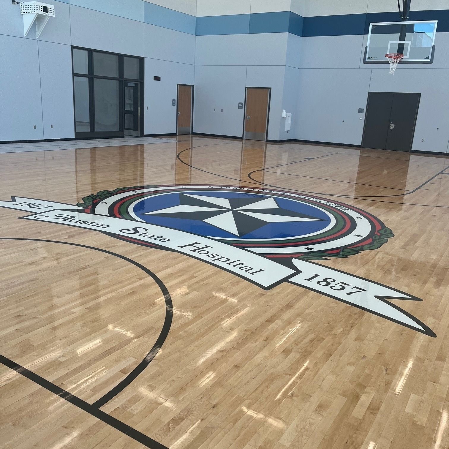 Empty gymnasium with polished wood floor and a large school emblem at center court.