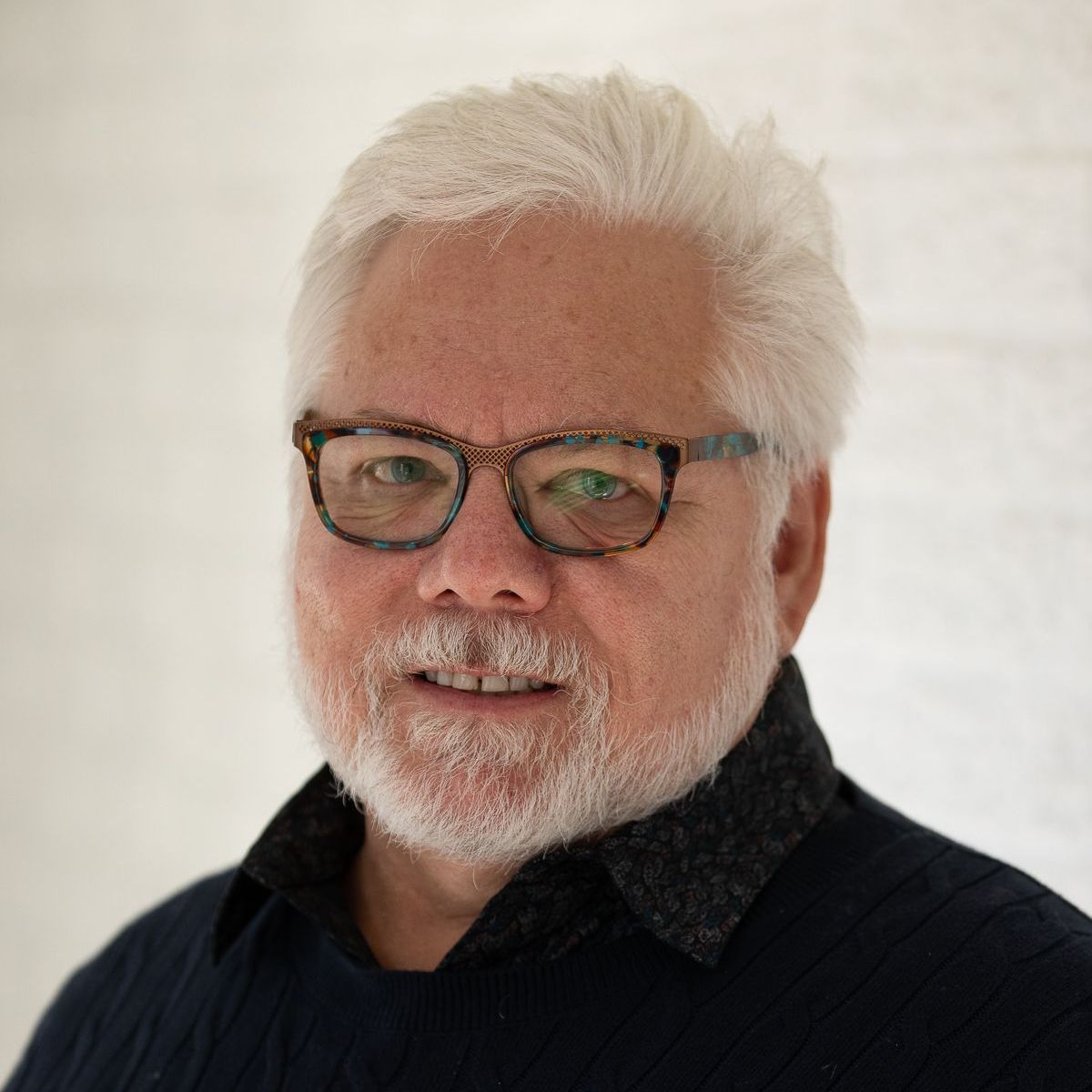 Man with glasses and white hair smiles, wearing a dark sweater against a white backdrop.