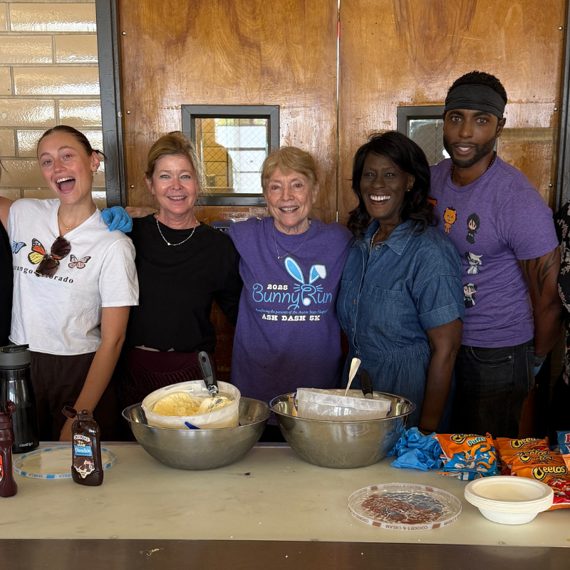 Five people smiling behind a table with bowls of food, in front of wooden doors.