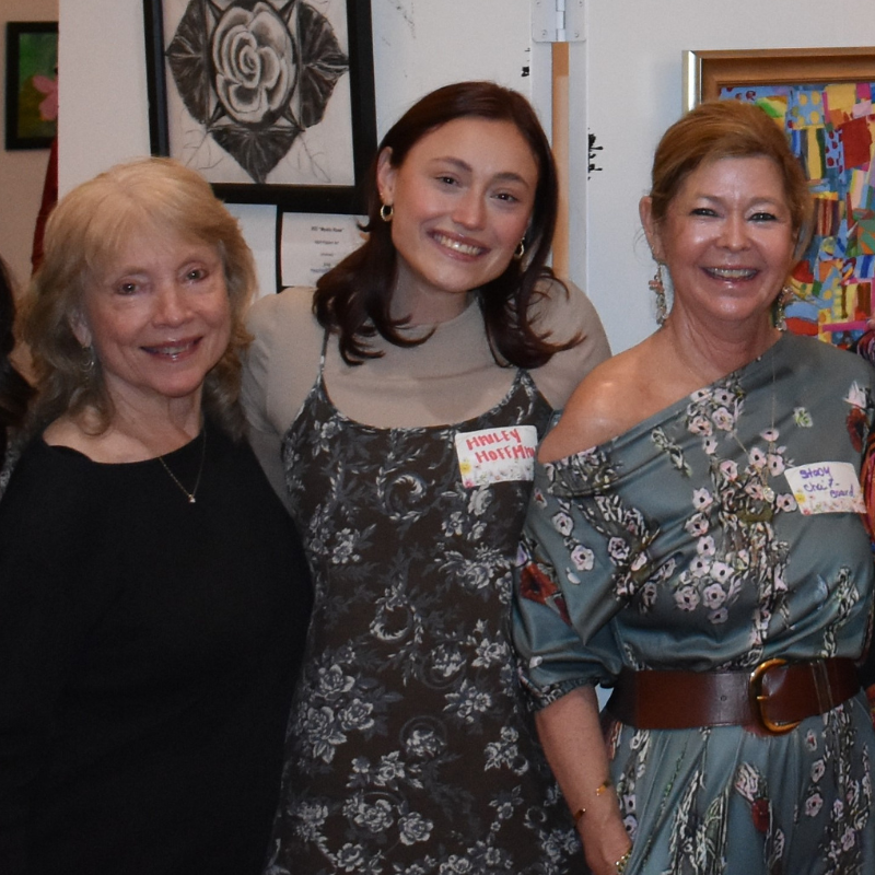 Three women smiling indoors at an art event, standing before framed artwork.