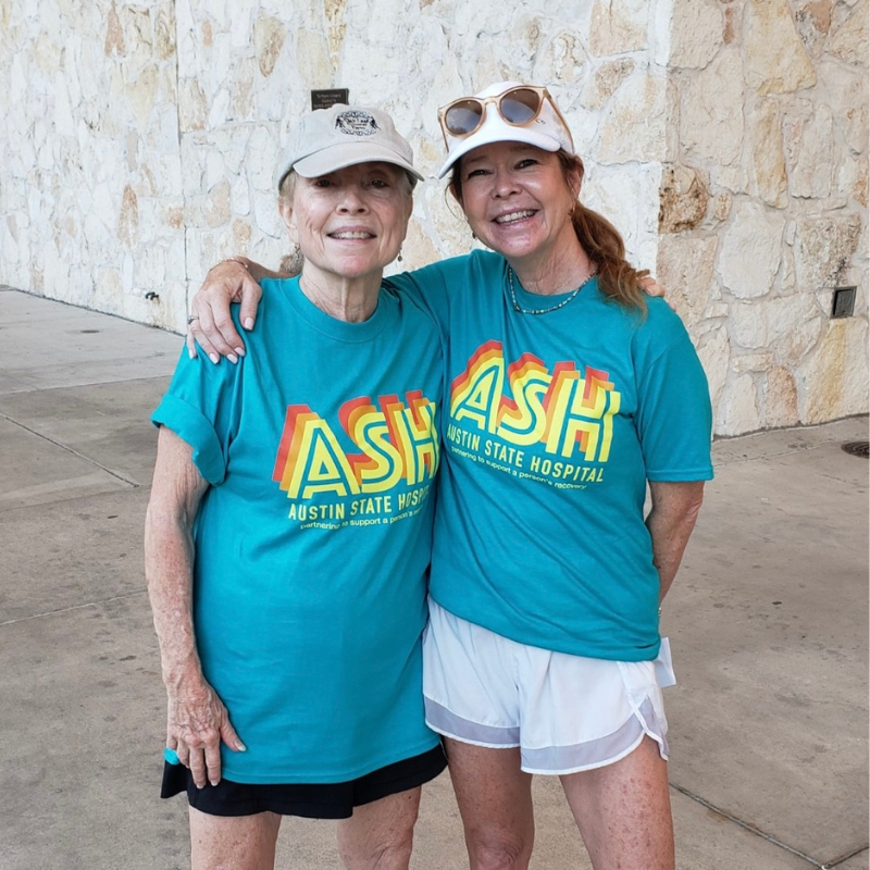 Two people in teal ASH shirts and white caps smiling outdoors against a stone wall
