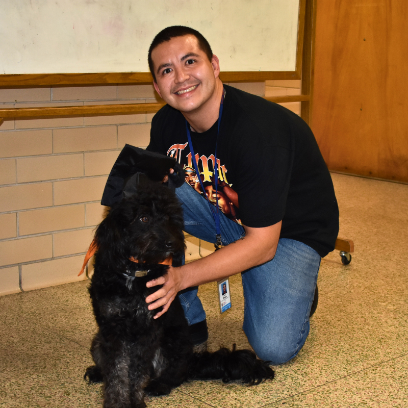Man kneeling indoors, smiling with a black dog beside him on a carpeted floor.