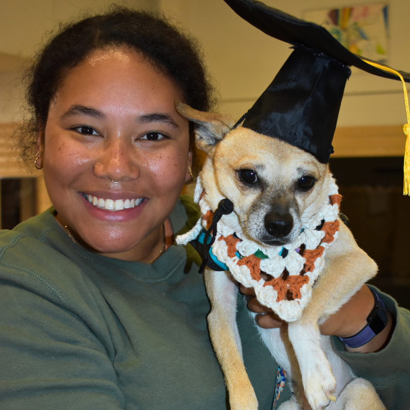 Smiling person holding a small dog in a graduation cap and lei outfit
