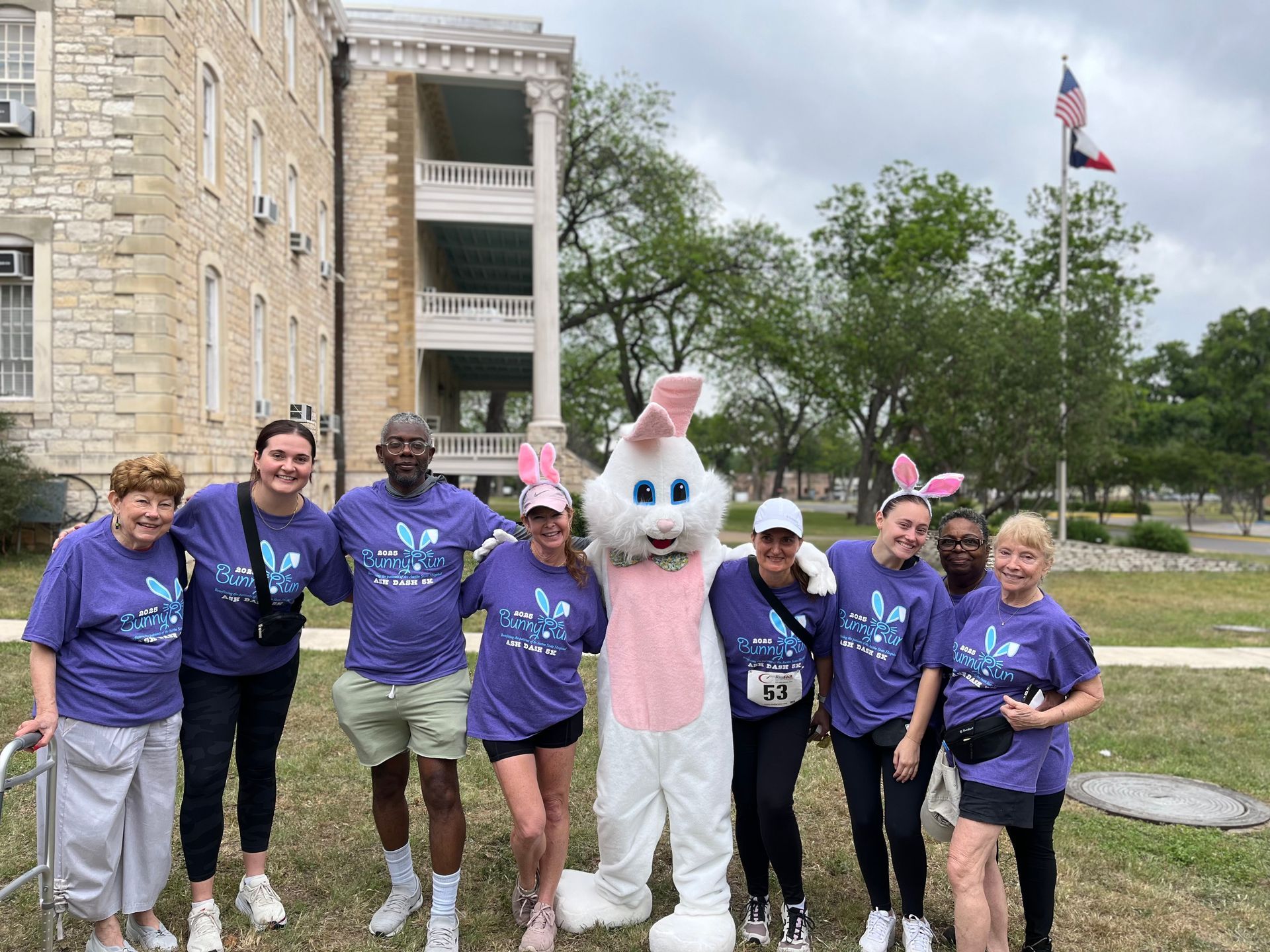 Group poses with a bunny mascot in front of a building and Texas flag; all wear purple shirts.