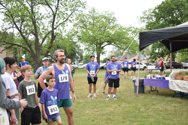Runners lined up at a grassy outdoor race start, with volunteers and a canopy tent nearby.