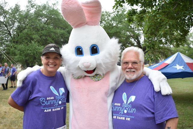 Two people in purple shirts pose with a pink-and-white bunny mascot outdoors by tents and trees.