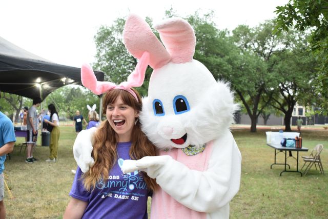 Woman posing with a white bunny mascot at an outdoor event, both smiling near tents and trees
