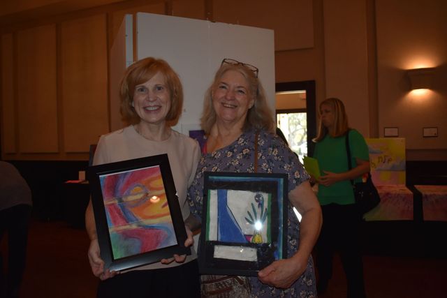 Two women holding framed artwork at an indoor event, smiling at the camera.