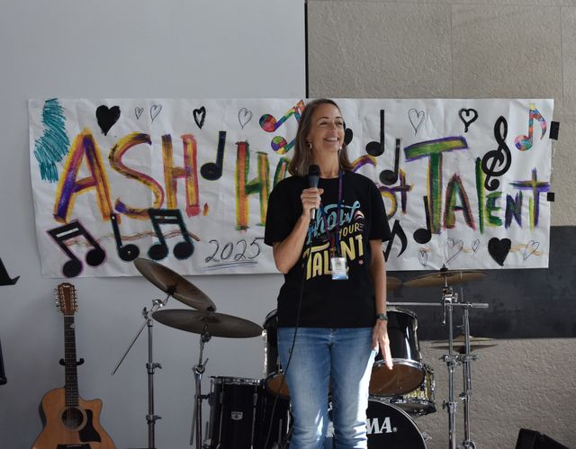 Person speaking on stage with drums, guitar, and a colorful “Ashes to…” banner behind them.