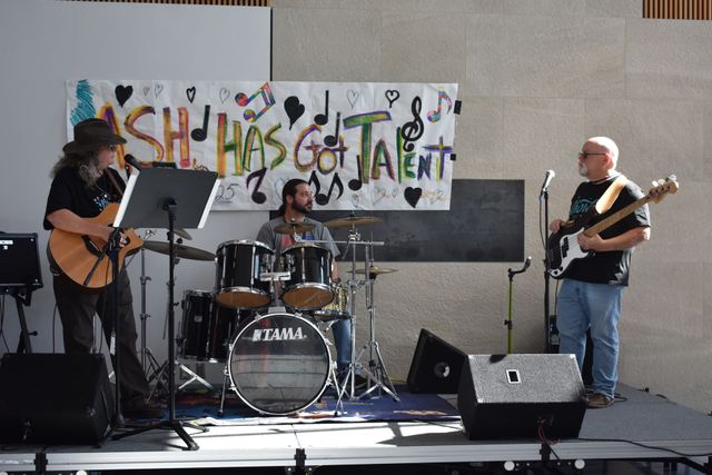 Three musicians perform on stage, with guitars and drums, in front of a “Fish Has Talent” banner.