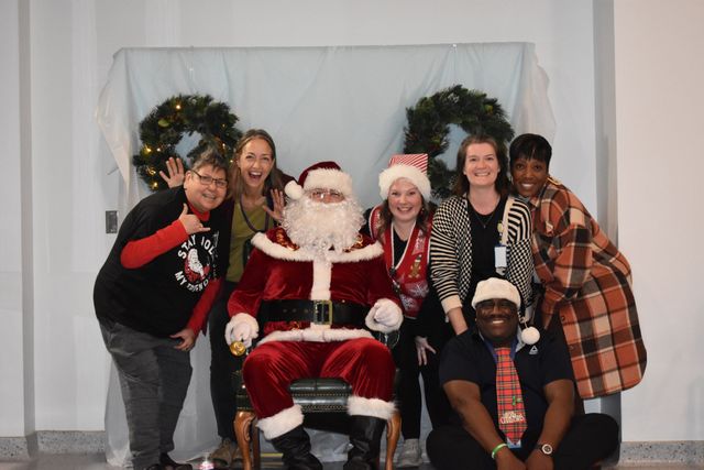 Group posing with Santa in front of holiday wreaths and white backdrop, smiling at the camera.