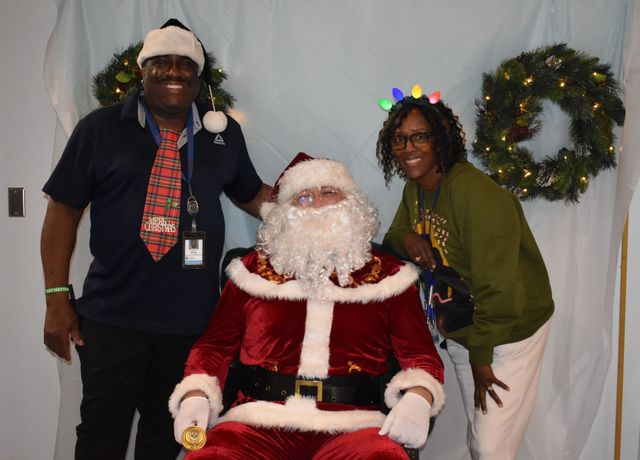Two people posing with Santa in front of holiday wreaths and a white backdrop.