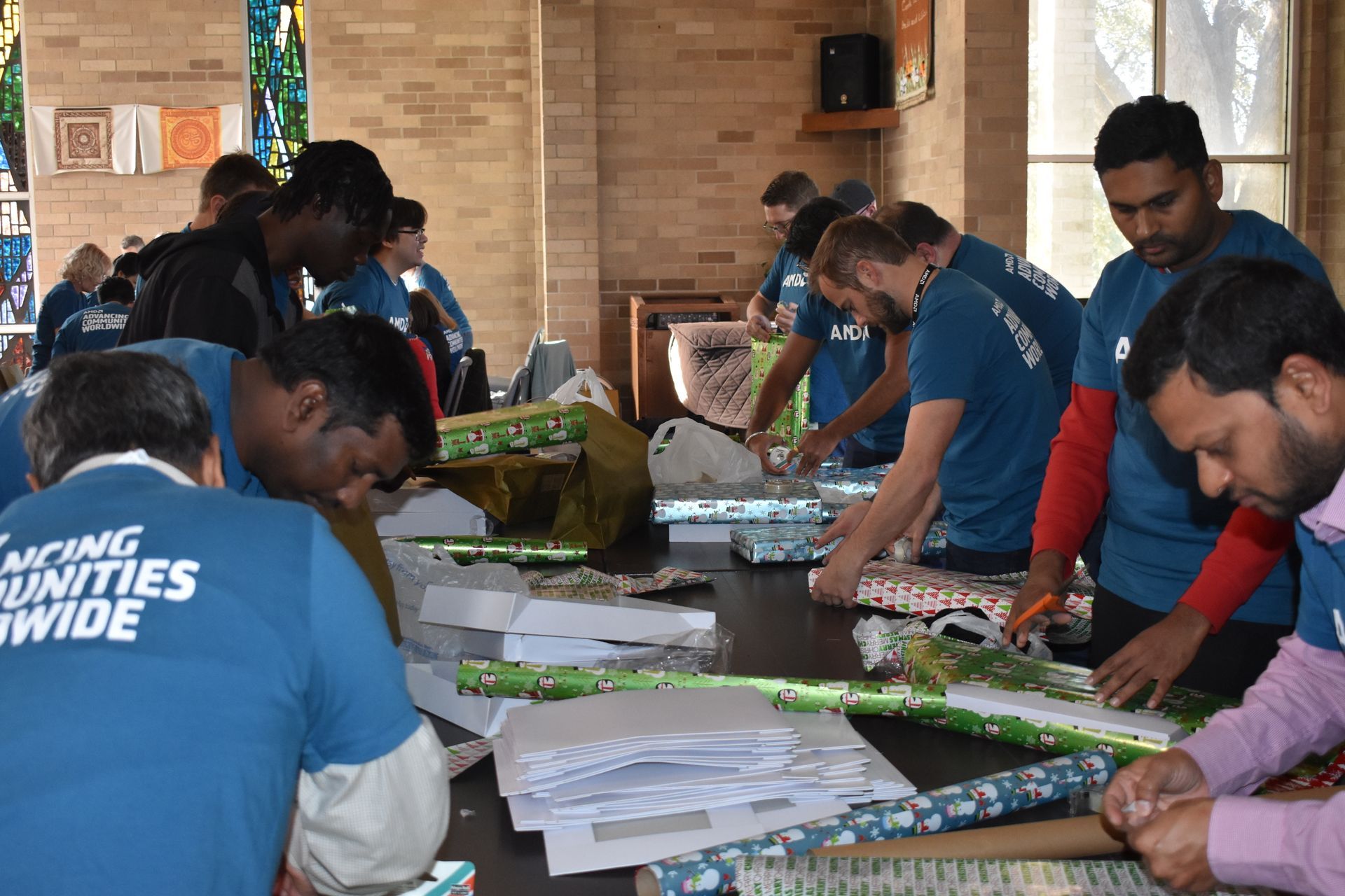 Volunteers in blue shirts assemble items at tables during a community event indoors