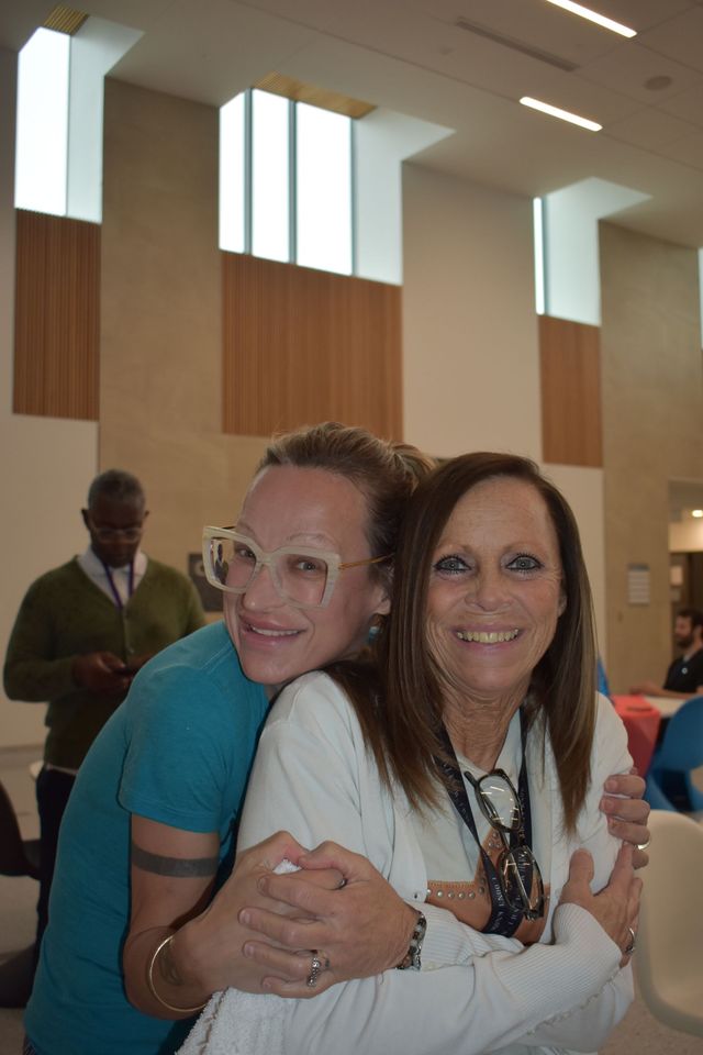 Two smiling women hugging indoors in a bright lobby with wood paneling