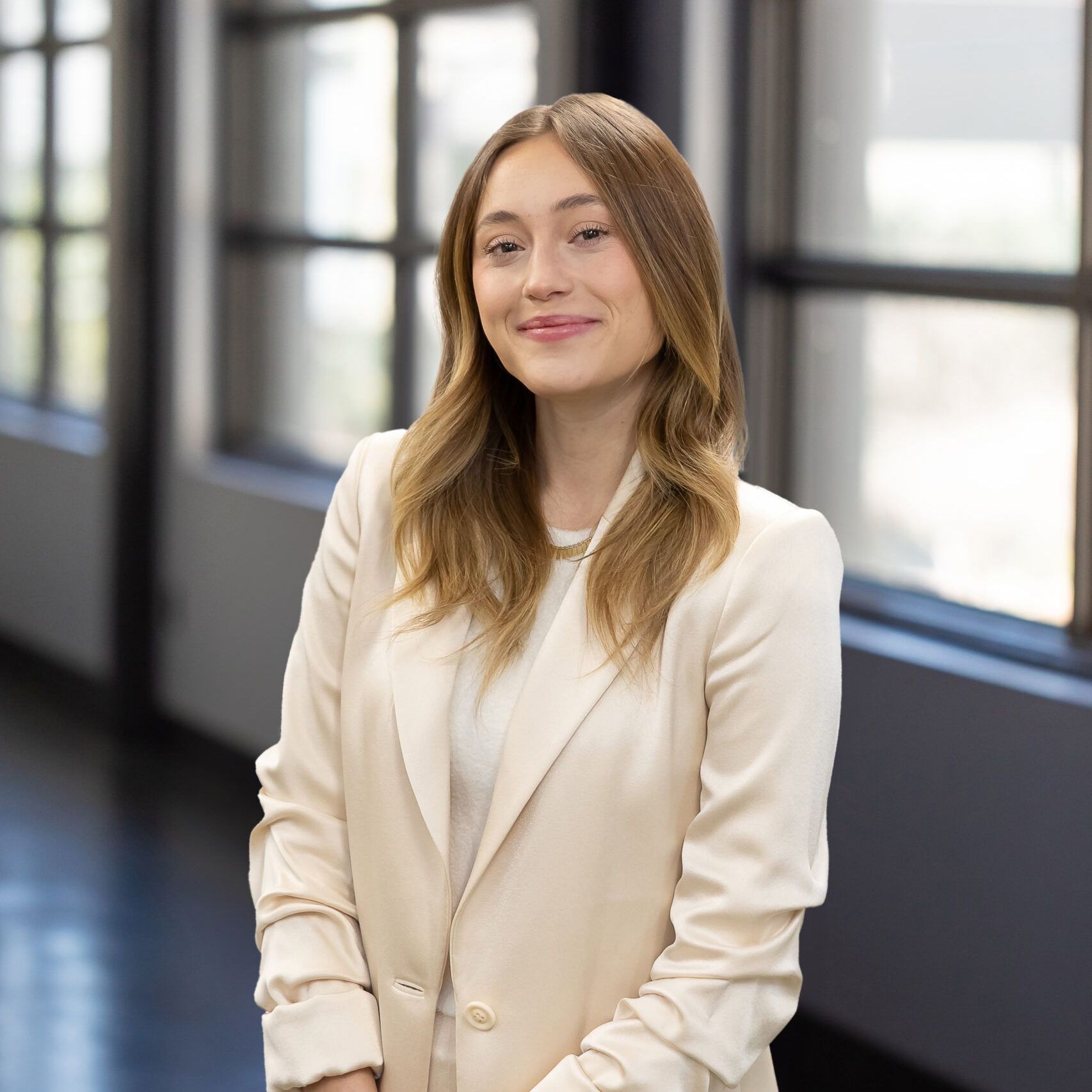 Woman in a light blazer smiles in a hallway with windows in the background.