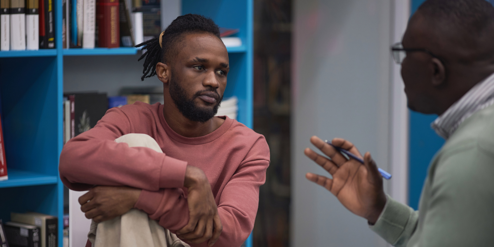 Two people talking in a room with shelves, one seated in a pink shirt listening attentively