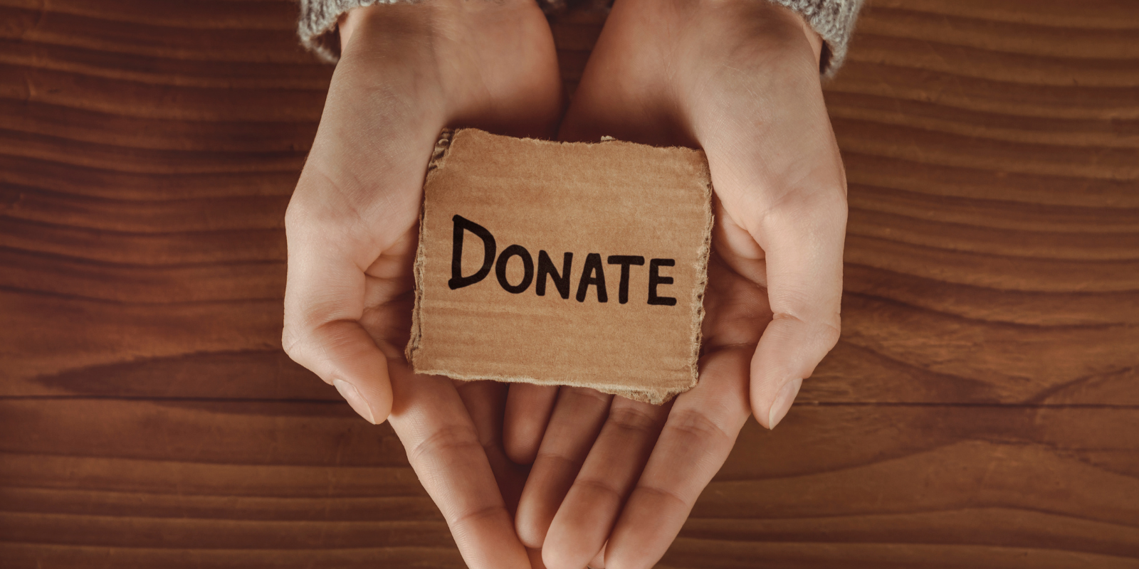 Hands holding a small cardboard sign labeled “DONATE” over a wooden background