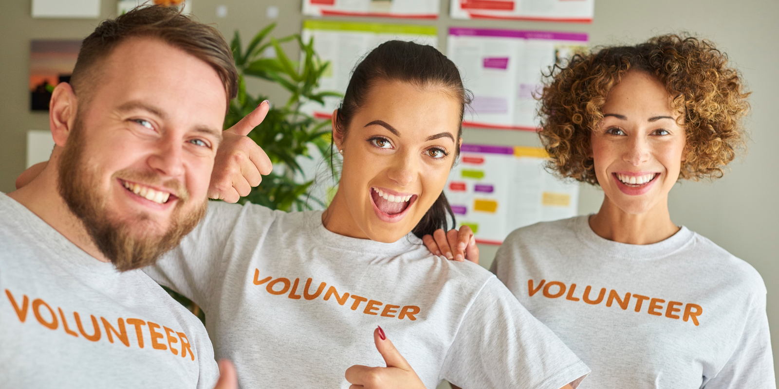 Three smiling volunteers in white shirts with orange “VOLUNTEER” text stand together indoors.