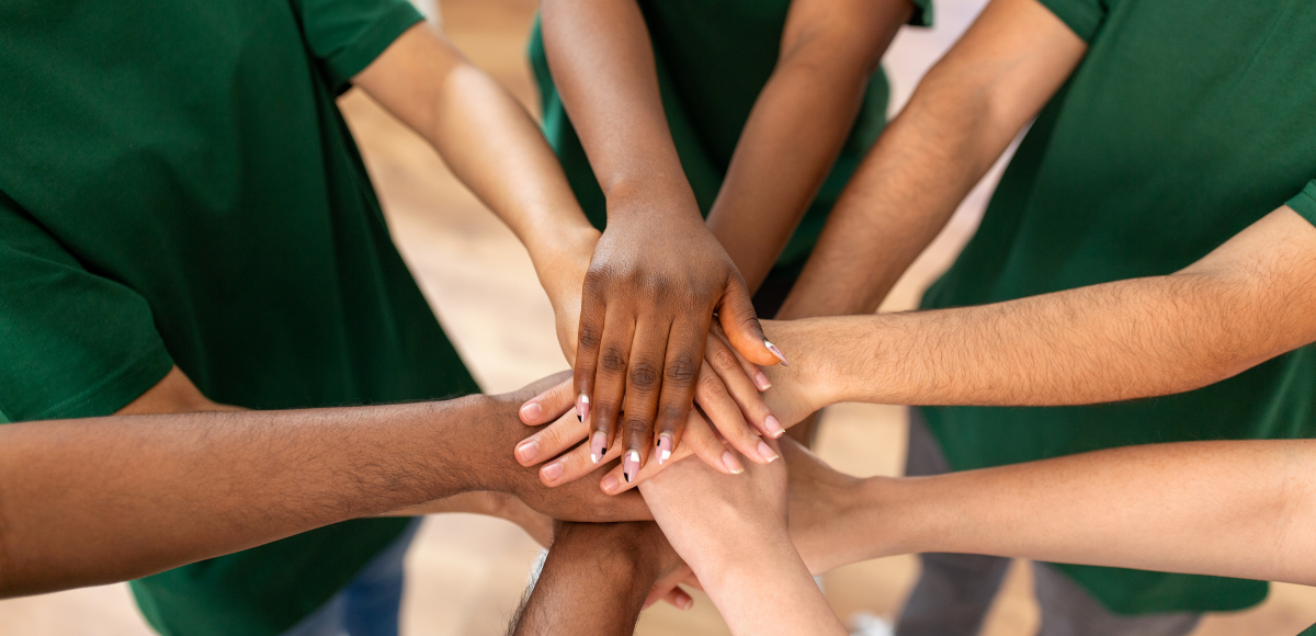 Stacked hands in a team huddle, with green shirts around the circle