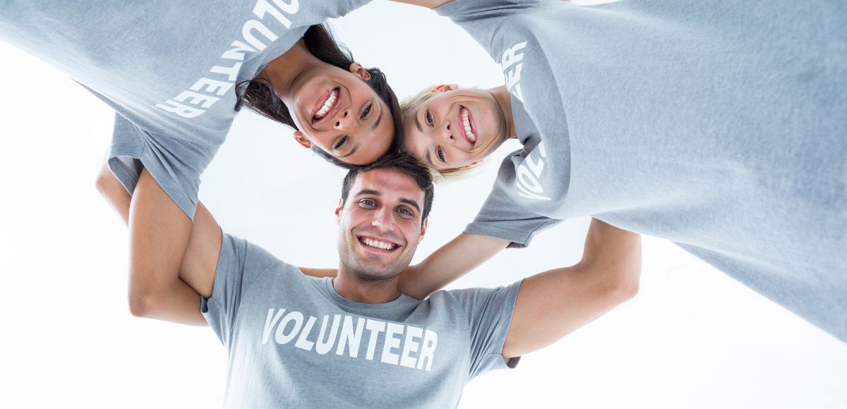 Three smiling volunteers in gray shirts huddle in a circle, viewed from below.