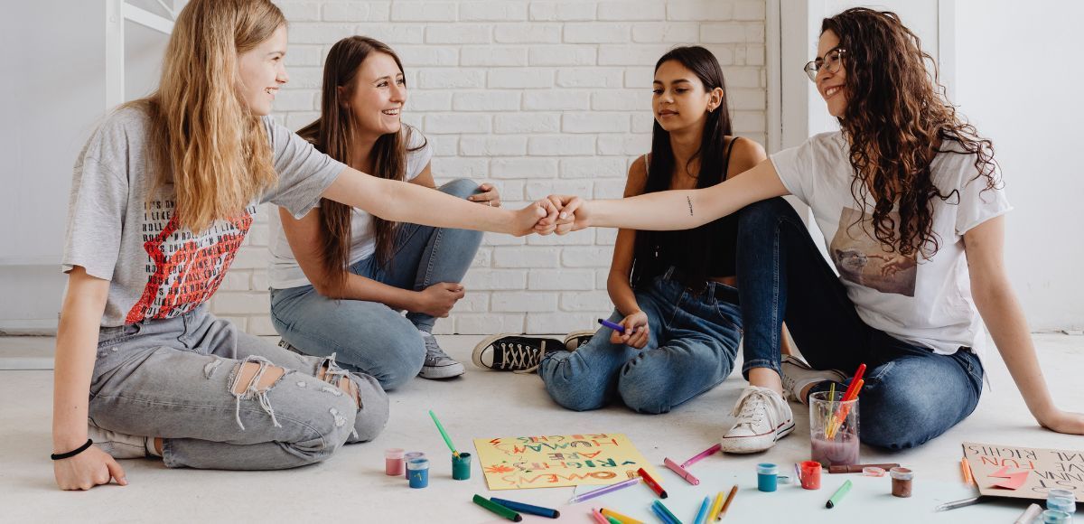 Four people sitting on the floor, reaching hands together in a creative craft session with art supplies.