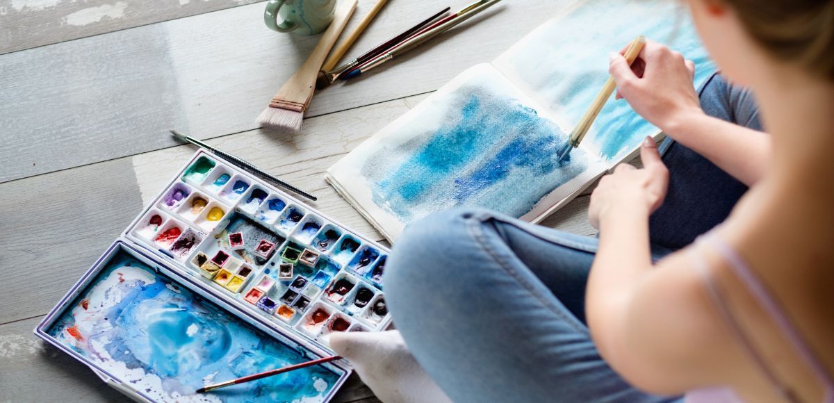 Person painting with blue watercolor at a table, with a palette and sketchbook nearby