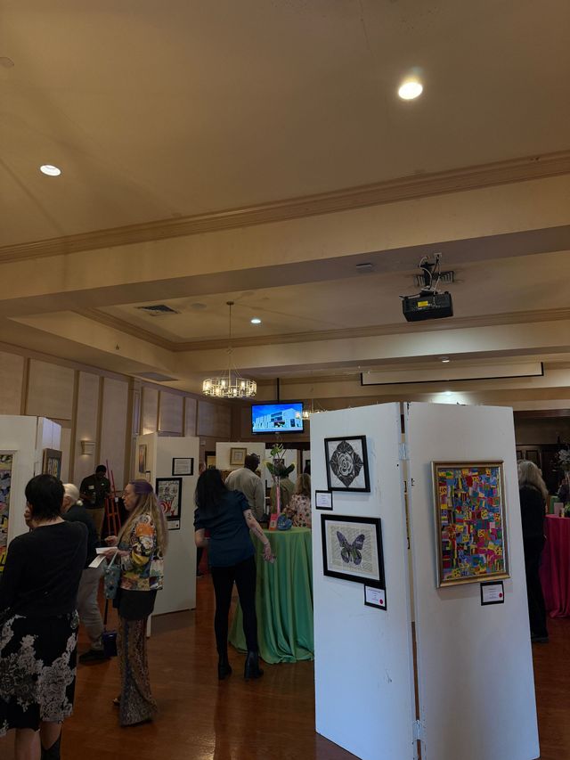 Art gallery interior with visitors, framed artwork on white panels, and a ceiling-mounted projector.