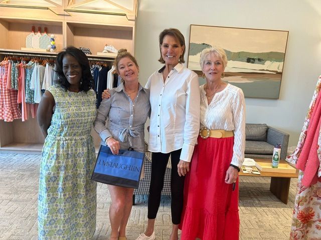 Four women standing together in a boutique, smiling and posing near clothing racks and a seating area.