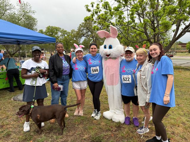 Group posing outdoors with a person in a bunny costume, blue shirts, and a dog at a charity event