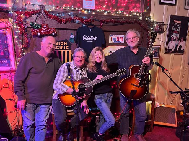 Four musicians posing with guitars on a colorful stage in a cozy bar setting